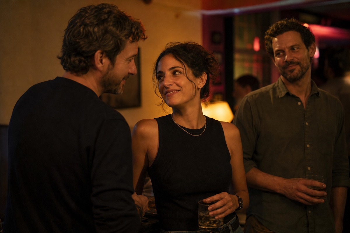 Three adults at a bar: a woman talks warmly with one man while another watches with an affectionate but thoughtful expression
